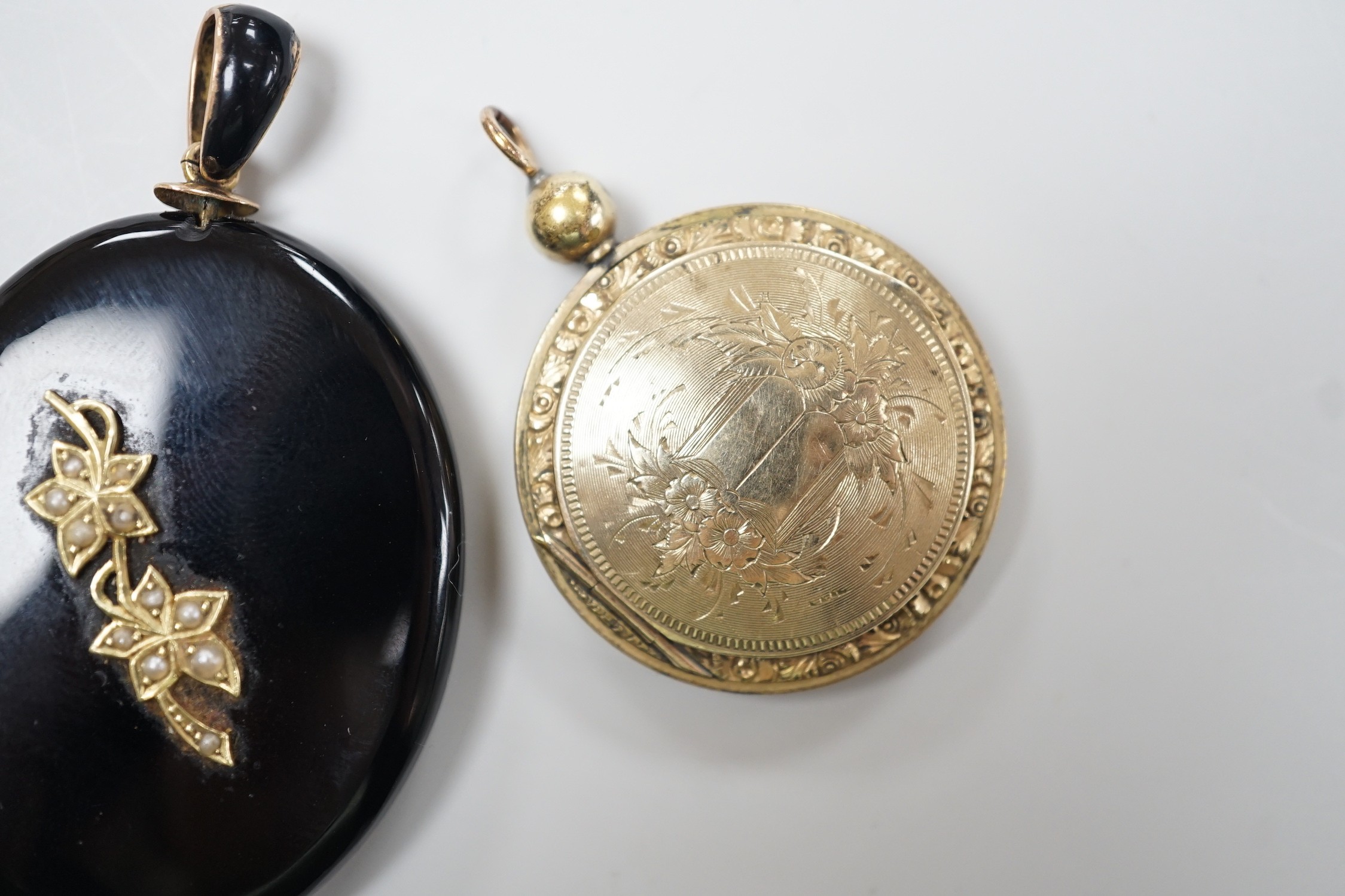A Victorian yellow metal and seed pearl mounted black onyx oval pendant locket, with enamelled bale, overall 53mm and an engraved gold plated circular pendant locket.